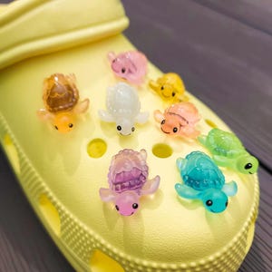 May include: A yellow pair of clogs with colourful turtle charms attached to the holes. The charms are made of translucent plastic and come in a variety of colours, including pink, blue, green, yellow, and orange.