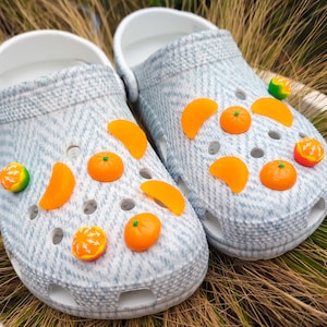 Può includere: Un paio di zoccoli azzurri decorati con ciondoli di frutta arancione e verde. I ciondoli includono arance intere, spicchi d'arancia e un frutto verde e giallo. Gli zoccoli hanno un cinturino bianco e un motivo strutturato.