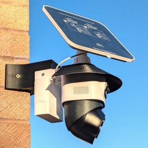 May include: A security camera system with a solar panel, mounted on a brick wall. The camera is black and white, with a black protective cover. The solar panel is angled towards the sky. The system is designed for outdoor use.