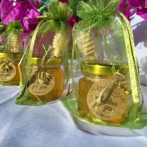 Pode incluir: Pequenos frascos de vidro cheios de mel, amarrados com barbante e um mergulhador de mel de madeira. Cada frasco tem uma etiqueta redonda de papel com o texto "With love for you". Os frascos são embalados em sacos de organza verde com uma fita.