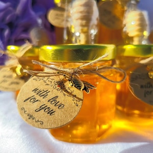 Peut inclure: Petits pots en verre remplis de miel, décorés de ficelle et d'un petit charme en forme d'abeille. Chaque pot est muni d'une étiquette en papier brun avec le texte "with love for you".