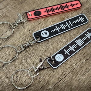 Könnte beinhalten: Drei Schlüsselanhänger mit Spotify-Code-Design. Einer ist korallenfarben, die anderen beiden sind schwarz und weiß. Jeder Schlüsselanhänger hat einen silberfarbenen Verschluss und einen Ring. Die Schlüsselanhänger sind rechteckig mit einem einzigartigen Barcode-Muster.