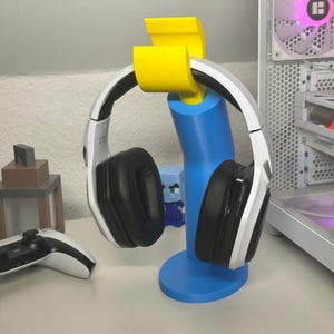 Könnte beinhalten: Ein weißes und schwarzes Gaming-Headset hängt an einem blau-gelben Kopfhörerständer. Ein weißer und schwarzer Gamecontroller liegt auf einer weißen Oberfläche. Ein Computerturm befindet sich im Hintergrund.