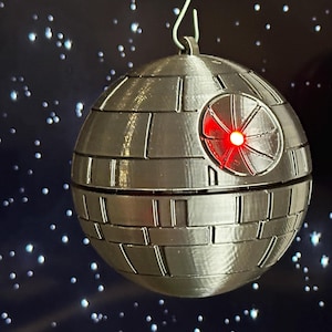 Könnte beinhalten: Grau-metallischer Death Star-Ornament mit rotem Licht, das von einem kreisförmigen Bereich ausgeht. Das kugelförmige Ornament hat eine detaillierte, paneelartige Oberfläche und wird von einem silbernen Haken gehalten. Der Hintergrund ist dunkelblau mit weißen sternartigen Punkten.