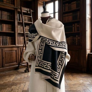 May include: A cream-coloured coat worn with a large, rectangular scarf featuring a black and cream Greek key pattern. The scarf is draped over the shoulders and chest, showcasing a bold geometric border design and a soft, fringed edge finish.