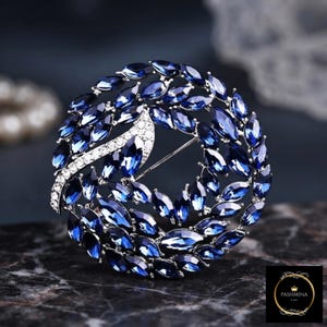 May include: A circular brooch featuring numerous marquise-cut deep blue crystals arranged in a wreath pattern. A curved silver-toned accent set with small clear rhinestones cuts across the left side. The logo PASHMINA is visible in the corner.