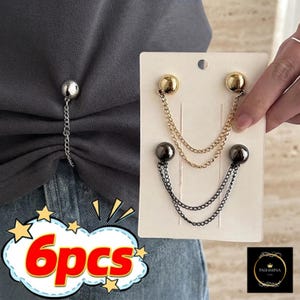 May include: A set of six metal clothing pins featuring spherical heads in silver, gold, and dark grey tones, each connected by two delicate link chains. The pins are designed to gather fabric, shown attached to a grey shirt and on a display card.