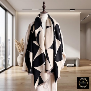 May include: A large, rectangular scarf featuring a bold, repeating black and cream geometric petal pattern. The scarf is draped over a mannequin wearing a cream-colored coat. It has a soft, woven texture and finished edges with short, delicate fringe.