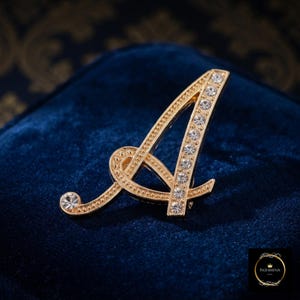 May include: A gold-toned metal brooch in the shape of the uppercase letter A. The letter features a textured, beaded border along one side and a row of clear, round, faceted rhinestones embedded along the other side, with a single rhinestone at the tip.