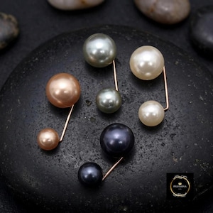 Könnte beinhalten: Eine Sammlung von fünf doppelseitigen Perlenstecknadeln mit Metallstielen und kugelförmigen Perlen in verschiedenen Farben wie Creme, Pfirsich, Grau und Schwarz. Jede Nadel besteht aus einer größeren und einer kleineren Perle an einem Draht.