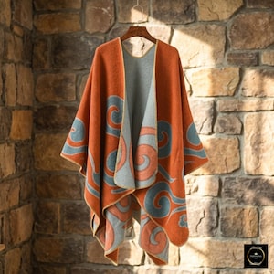 May include: A reversible open-front poncho featuring a burnt orange exterior with large, swirling blue patterns and a contrasting light blue interior with burnt orange swirls. The garment has finished edges and is displayed on a wooden hanger.