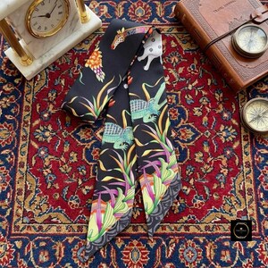 May include: A long, narrow black scarf featuring a vibrant, multi-coloured print of various animals, including a giraffe, elephant, and crocodile, interspersed with tropical foliage in shades of green, yellow, and purple on a dark background.