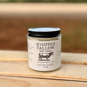 Könnte beinhalten: Ein Glas mit geschlagener Talg-Körpercreme mit schwarzem Deckel. Das Etikett zeigt "Whipped Tallow Body Cream" mit einer Kuh-Illustration und "Candy Cane 4 oz". Das Glas steht auf einer Holzoberfläche.
