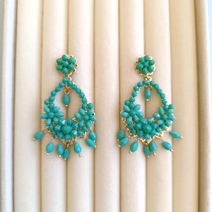 Puede incluir: Un par de pendientes colgantes turquesa con detalles dorados. Los pendientes presentan un diseño floral en la parte superior y cuentas en cascada en forma de lágrima. Los pendientes se exhiben sobre una superficie color crema.