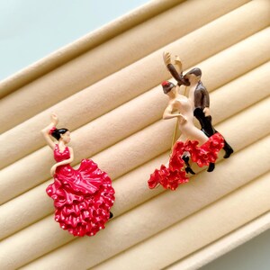 Peut inclure: Deux figurines de danseurs de flamenco. Une danseuse porte une robe rouge avec un accessoire pour les cheveux noir. L'autre figurine montre un couple dansant, la femme en robe rouge et l'homme en costume. Les figurines sont sur une surface beige.