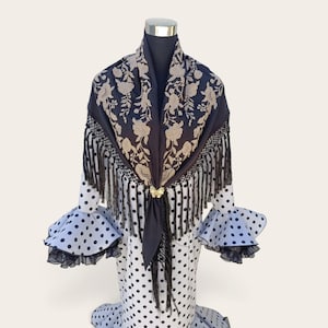Peut inclure: Un châle triangulaire noir avec des broderies florales beiges et de longues franges noires, fixé par une broche papillon dorée. Il est posé sur une robe blanche à pois noirs avec des manches à volants bordées de finitions noires.