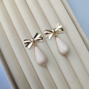 May include: A pair of gold-tone bow earrings featuring a pale pink, teardrop-shaped bead. The earrings are presented in a cream-coloured jewellery box with a velvet lining. Each earring has a small gold bead at the base of the teardrop.