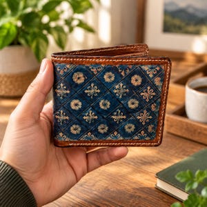 Puede incluir: Una cartera plegable con un exterior azul profundo y un patrón de diamantes acolchados. Cada diamante contiene un pequeño y complejo motivo floral en tonos dorados y bronceados. La cartera está rematada con un borde de cuero marrón oscuro.