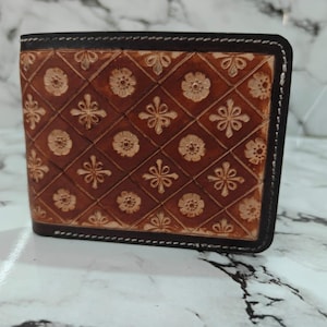 Puede incluir: Una cartera plegable de cuero color canela con un borde marrón oscuro y costuras blancas. La superficie está grabada con un patrón de diamantes repetitivo, y cada diamante contiene un pequeño y detallado diseño floral en el centro.