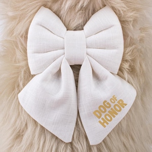 Op de afbeelding: Witte stoffen strik met de tekst "DOG OF HONOR" geborduurd in goud.