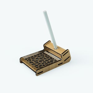 May include: A wooden pencil holder with a geometric pattern on the base and a slot for a pencil. The pencil holder is made of light brown wood and has a white pencil inserted in the slot.