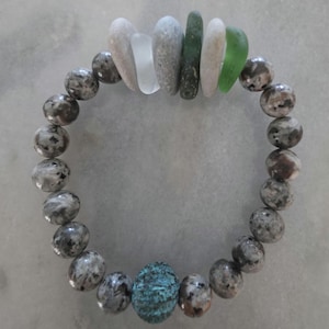 Puede incluir: Una pulsera elástica con cuentas redondas de piedra gris y marrón moteada. El punto focal incluye una cuenta texturizada de color verde azulado en la parte inferior y un grupo de piezas de vidrio marino lisas, planas, blancas, grises y verdes arriba.