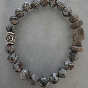 Può includere: Un braccialetto elastico con perline rotonde in pietra maculata grigia e marrone. Il design include una perlina distanziatrice in metallo argentato con un motivo a spirale e un ciondolo in pietra marrone chiaro intagliato a forma di pesce.