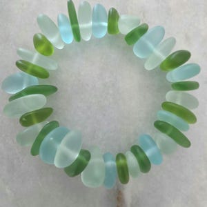 Peut inclure: Un bracelet circulaire composé de morceaux de verre de mer dépoli et lisse, alternant des nuances de bleu clair, de vert menthe et de vert citron. Les segments de verre individuels sont arrondis et polis, assemblés en une boucle continue.