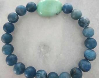 May include: A beaded bracelet featuring round, blue beads and a central, oval-shaped, light green stone. The bracelet is made of natural stones and is designed to be worn around the wrist. The beads have a matte finish.