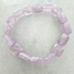 May include: A bracelet made of light purple, rectangular amethyst beads. The beads are strung together to form a circular shape. The bracelet is displayed on a light gray surface.