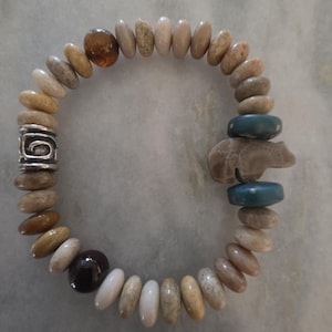 Puede incluir: Una pulsera elástica con una serie de cuentas de piedra lisas y redondas en tonos tierra, incluyendo beige, bronceado y marrón. La pulsera incluye un espaciador de metal plateado con un diseño en espiral, dos cuentas de disco azules y un dije.