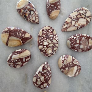 May include: A collection of nine polished cabochons featuring a brecciated pattern with a mix of deep maroon, reddish-brown, and creamy beige angular fragments. The stones are shaped into various ovals and teardrops, displaying a smooth, glossy finish.