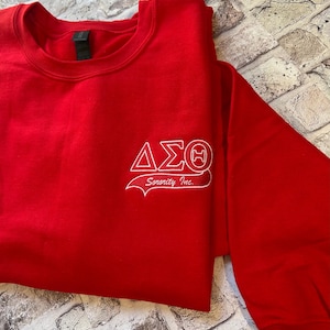 May include: A bright red long-sleeve crewneck sweatshirt featuring white embroidery on the upper left chest. The embroidery displays the Greek letters Delta Sigma Theta and the text Sorority Inc. in a cursive script font below the letters.