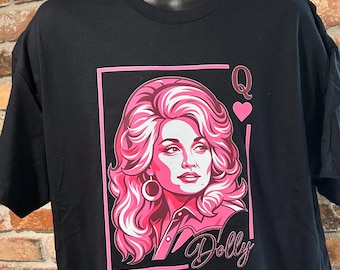 May include: A black short-sleeve t-shirt featuring a graphic print of a woman with voluminous pink hair styled as a playing card queen. The design includes the letter Q, a pink heart, and the script text Dolly in pink and white ink colors.