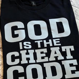 May include: A black short-sleeved t-shirt featuring the text "GOD IS THE CHEAT CODE" printed in large, bold, light gray capital letters across the front. The shirt has a standard crew neckline and a visible brand label at the inner collar.
