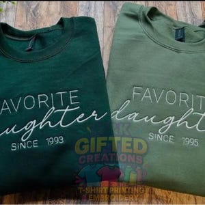 Puede incluir: Dos sudaderas verdes con el texto "FAVORITE daughter" bordado en blanco. Una es verde oscuro, la otra verde oliva. La verde oscuro dice "SINCE 1993" y la verde oliva "SINCE 1995".