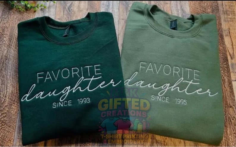 Puede incluir: Dos sudaderas verdes con el texto "FAVORITE daughter" bordado en blanco. Una es verde oscuro, la otra verde oliva. La verde oscuro dice "SINCE 1993" y la verde oliva "SINCE 1995".