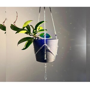 May include: A hanging planter with a dark grey pot, filled with green leafy plants. The pot is suspended by beaded chains and a decorative crystal pendant. The plant pot is approximately 15 cm in diameter.