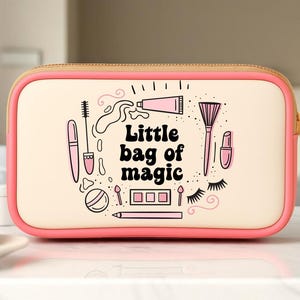 May include: A rectangular makeup bag with a pink border and a beige center. The bag features illustrations of makeup items and the text "Little bag of magic". A gold zipper and a white cup are also visible.