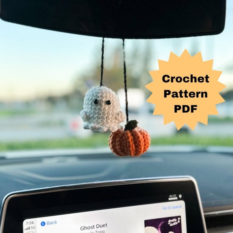 May include: Crocheted Halloween car charm featuring a white ghost and an orange pumpkin with a green stem. The charm is suspended by black strings. The image also includes text that says "Crochet Pattern PDF".
