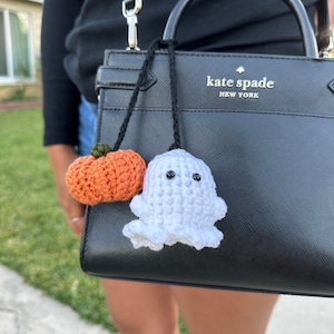 May include: Crocheted Halloween-themed bag charms. One charm is an orange pumpkin with a green stem, and the other is a white ghost with black eyes. Both charms are attached to a black cord.