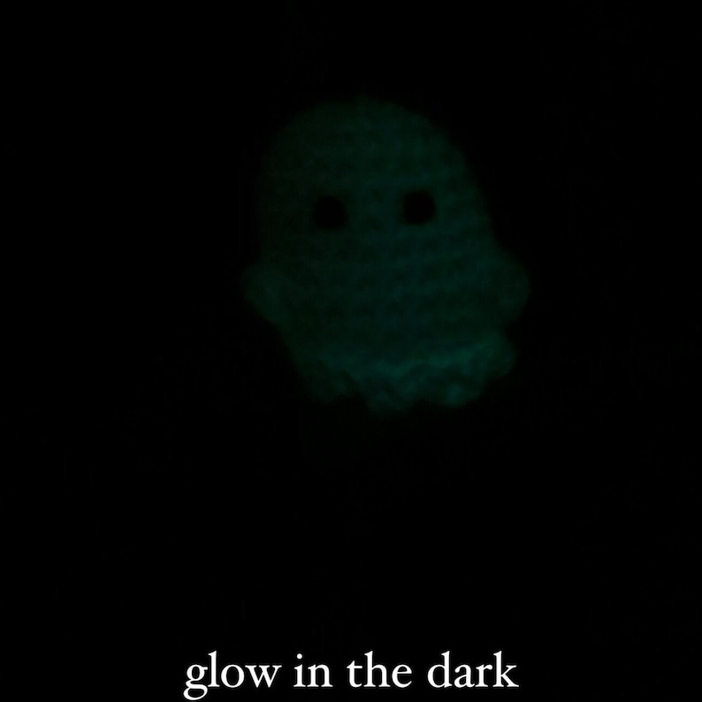 May include: A glowing, crochet ghost with two black eyes against a black background. The text "glow in the dark" is at the bottom of the image. The ghost is a light green color.