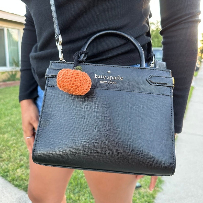 May include: Black Kate Spade handbag with a small, orange crocheted pumpkin charm. The charm is attached to the handle of the purse. The handbag has a top handle and a shoulder strap. The text "kate spade NEW YORK" is visible on the front of the bag.
