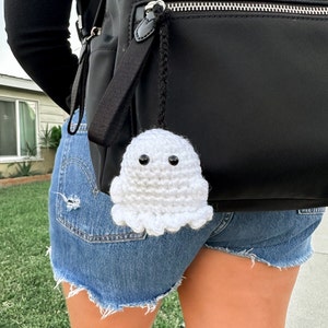 May include: A white crocheted ghost charm with black button eyes hangs from a black backpack. The ghost has a rounded body and a ruffled bottom edge. The charm is attached to the backpack with a black cord.