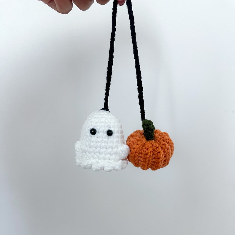 May include: Crocheted Halloween ornaments featuring a white ghost with black eyes and an orange pumpkin with a green stem, suspended by black cords. These handmade decorations are perfect for adding a festive touch to any space.