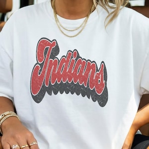 May include: A white short-sleeved t-shirt featuring the word "Indians" printed in a red, distressed, retro-style font with a thick black drop shadow. The graphic is centered on the chest of the garment, which has a classic crew neckline.