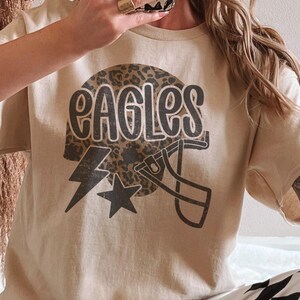 May include: A beige t-shirt featuring a football helmet design with the word "EAGLES" in a bold, black font. The helmet has a leopard print pattern. A black lightning bolt and star are also included in the design.
