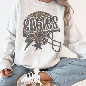 May include: A light gray sweatshirt with the word "EAGLES" in a distressed font, a football helmet, a lightning bolt, and a star graphic. The word "EAGLES" is set against a leopard print background.