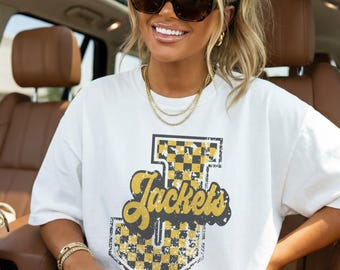 May include: A white short-sleeved t-shirt featuring a large graphic print on the front. The design includes a yellow and black checkered letter T with the word Jackets written in a cursive yellow font across the center, all with a distressed texture.