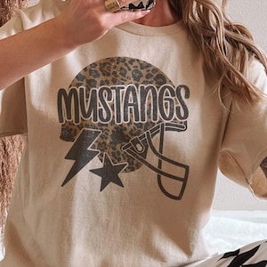 May include: A tan-colored t-shirt featuring a graphic design. The design includes the word "MUSTANGS" in a bold, dark font, a football helmet, a lightning bolt, and a star. The helmet has a leopard print pattern.
